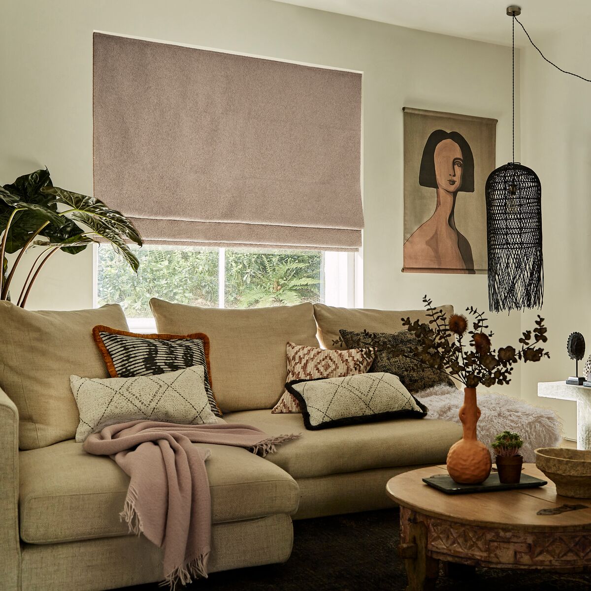 Taupe Roman blind above a bright window in a cosy living room, with a neutral sofa, patterned cushions, wooden coffee table and fireplace.