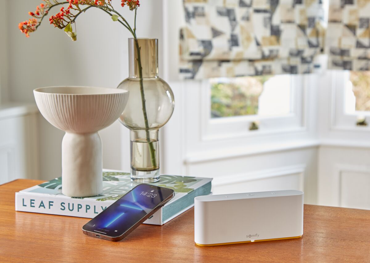 Patterned Roman blinds frame a bright corner window, with a smart home hub, phone and decorative vase on a wooden table.