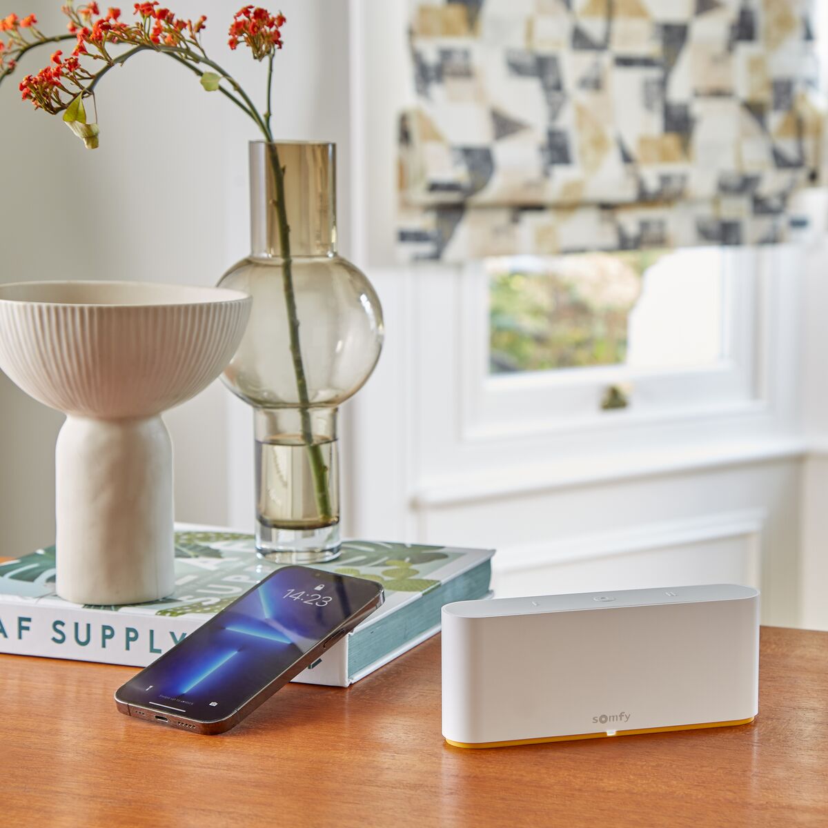 Patterned Roman blinds frame a bright corner window, with a smart home hub, phone and decorative vase on a wooden table.