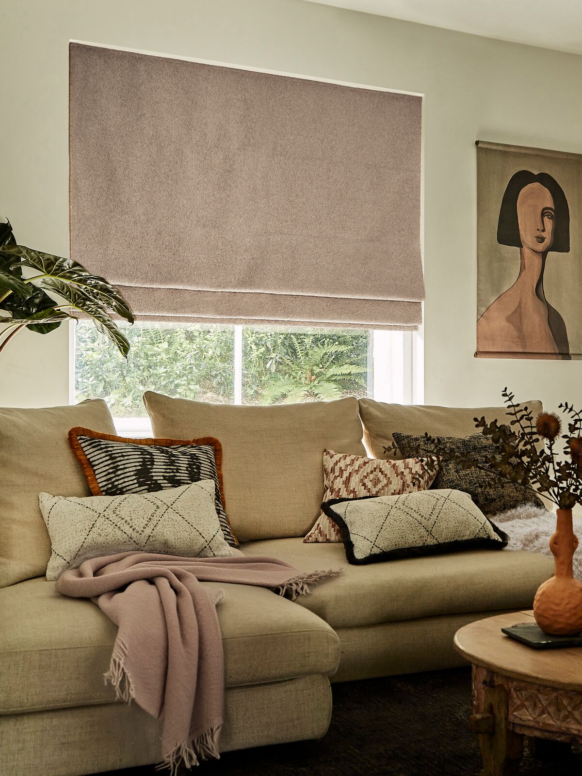 Taupe Roman blind above a bright window in a cosy living room, with a neutral sofa, patterned cushions, wooden coffee table and fireplace.
