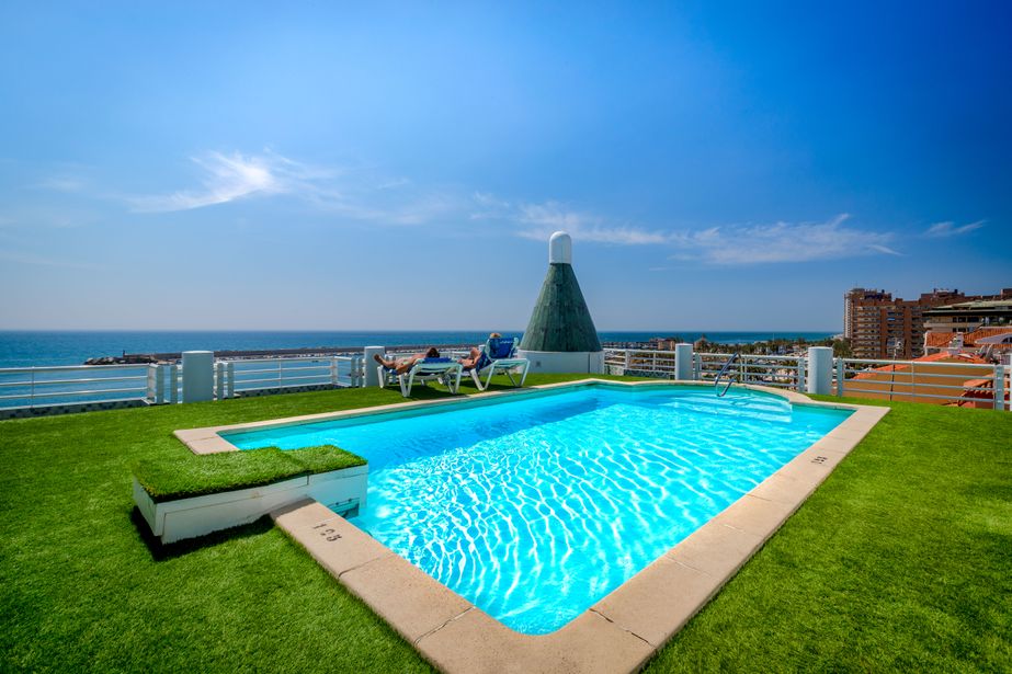 Rooftop swimming pool with turquoise water, sun loungers and sunbathers overlooking the sea and coastal skyline on a sunny day