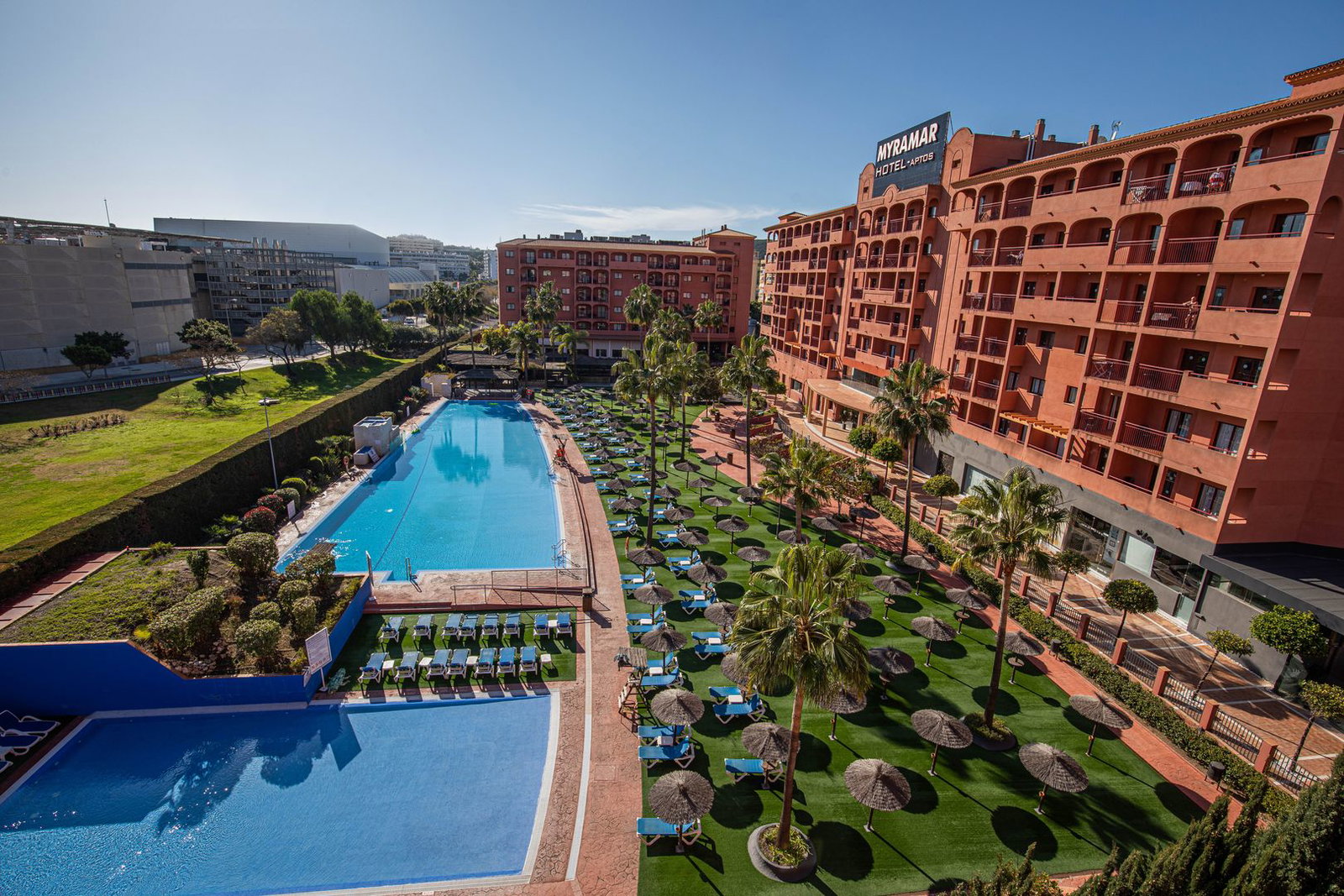 Aerial view of Myramar apartments with large outdoor swimming pools, sun loungers, palm trees and terracotta hotel façade