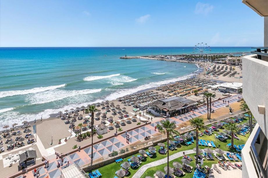 Elevated view of a busy Mediterranean beachfront with rows of sunbeds, promenade, turquoise sea and distant Ferris wheel