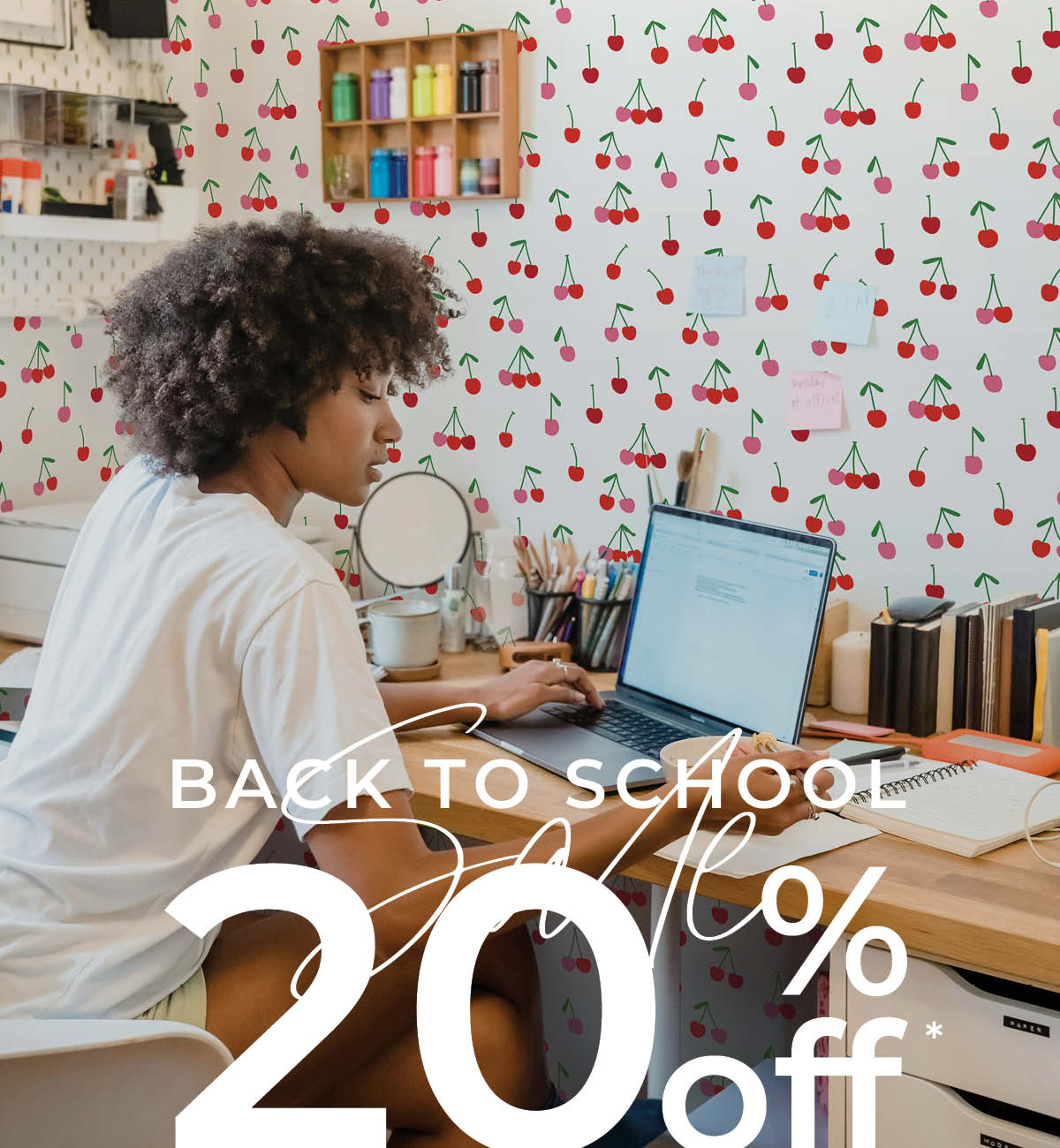 Image of Tempaper's Cherry Bomb wallpaper on a wall behind a student sitting at a desk. Text overlay "Back to School Sale 20% off*"