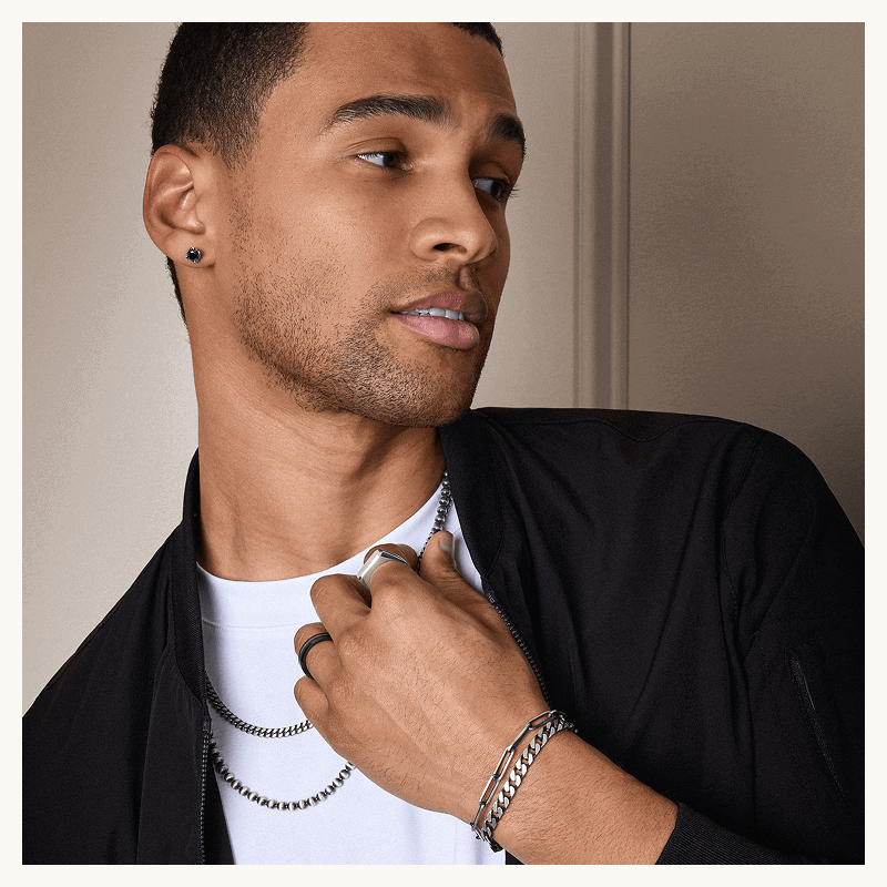 Male model in various silver & beaded chain bracelets & necklaces & black gemstone studs.