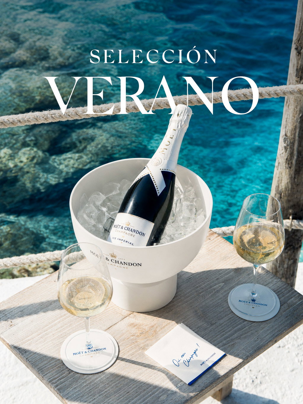 Bottle of champagne in an ice bucket with two filled wine glasses on a wooden deck overlooking turquoise sea in a summer setting