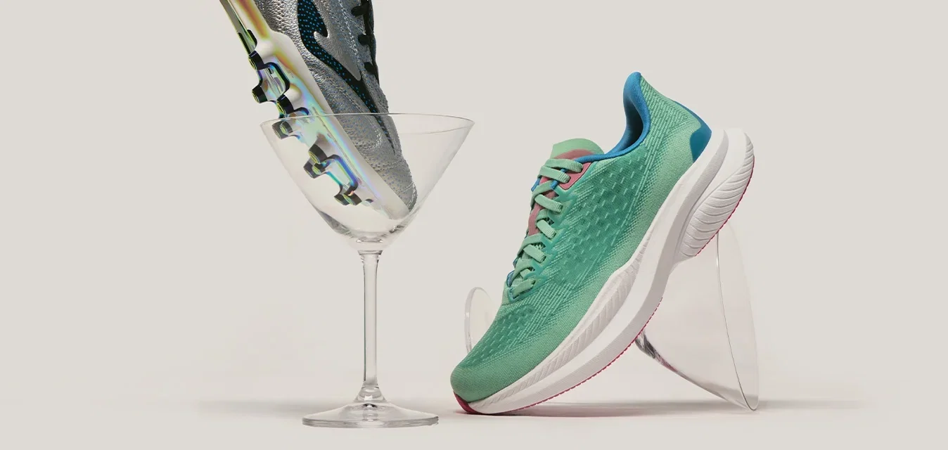 Botín de fútbol plateado balanceado en una copa de martini junto a un zapato deportivo verde inclinado sobre un soporte transparente en un fondo liso.