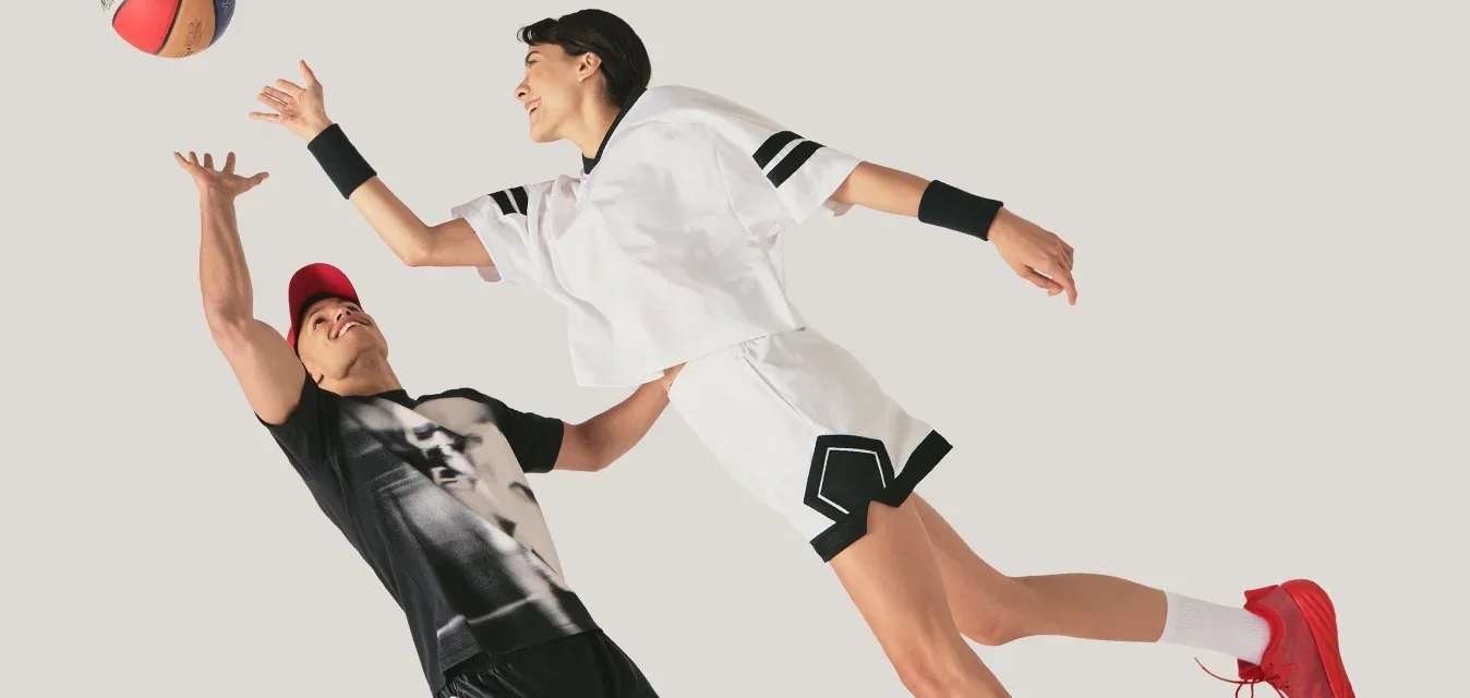 Dos jugadores de baloncesto en el aire alcanzando el balón, vestidos con ropa deportiva negra y blanca sobre un fondo liso.