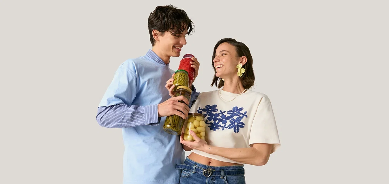 Joven hombre y mujer sonrientes, sosteniendo frascos de verduras en escabeche, posan frente a un fondo sencillo y claro.