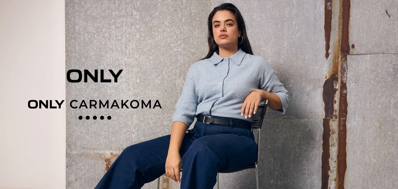 Mujer con el cabello largo sentada en una silla, lleva un jersey azul claro, pantalones azul oscuro y mocasines marrones, delante de una pared industrial gris.