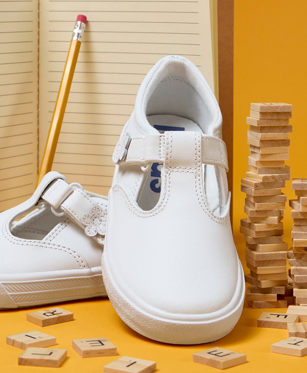 A pair of Little Kids’ Daphne T-Strap sneakers in white sitting in front of an open notebook showing blank lined paper. A wooden pencil is balanced against the notebook, and a stack of Scrabble letter tiles are to the right of the sneakers.