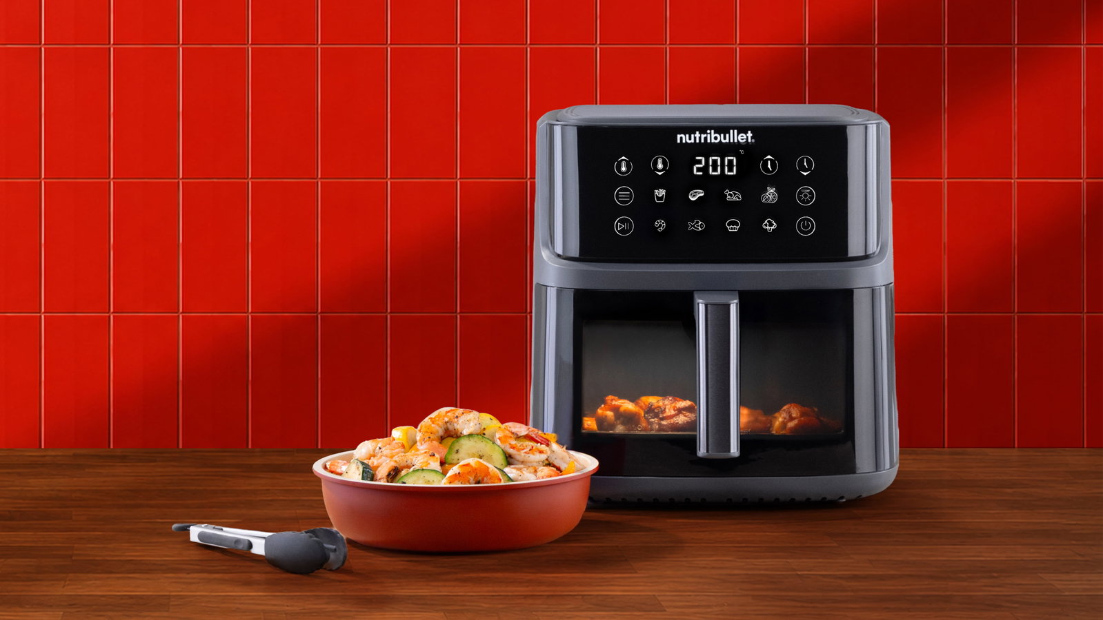A dark grey nutribullet CrispLite™ Vision Air Fryer displaying 200°C beside a ceramic bowl of cooked shrimp and vegetables.
