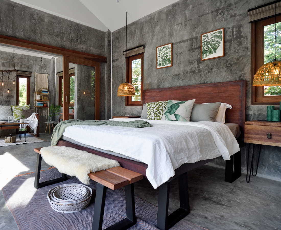 Image description: Boho hotel room with slate gray walls and flooring and dark wooden accents. There is a large bed in the center of the room with sliding doors that open up to a cozy living area with a couch and coffee table.