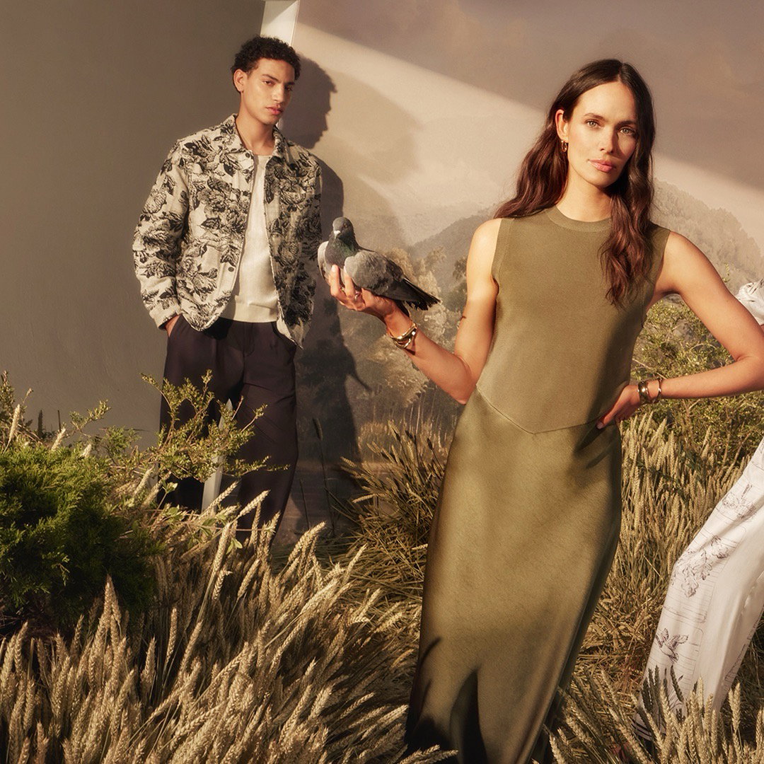 Woman in olive knit dress with a bird on her hand, man in printed jacket and navy trousers in background