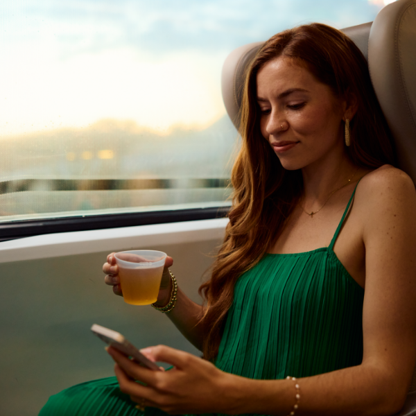 A young woman in a green dress sits by a large window on a train, holding a smartphone in one hand and a plastic cup with a light-colored beverage in the other. Soft daylight filters through the window, suggesting travel during the day. Her relaxed posture and slight smile convey a calm, peaceful mood. Neutral tones of the train interior contrast with the vibrant green of her dress.