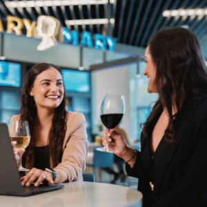 Two women sit at a round table in a contemporary indoor lounge, smiling and talking while holding glasses of wine. One woman works on a laptop with a glass of white wine, while the other raises a glass of red wine in a friendly toast. The background features bright signage and large digital screens, with a ceiling of linear lights adding a modern feel. The overall atmosphere is social, professional, and relaxed.