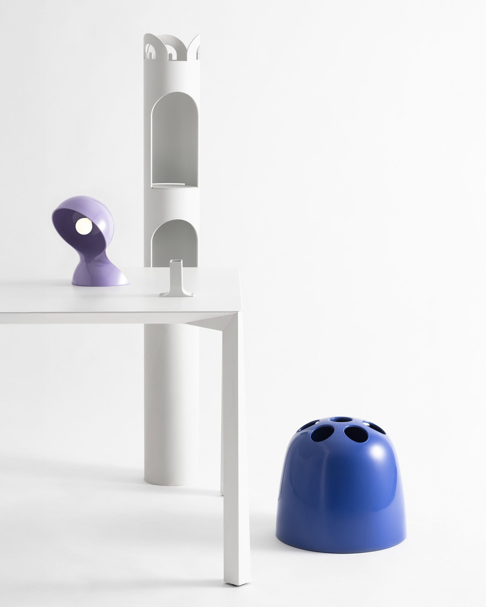 Minimalist white interior with a white table, purple modern desk lamp, tall white shelf and a blue rounded stool with circular holes