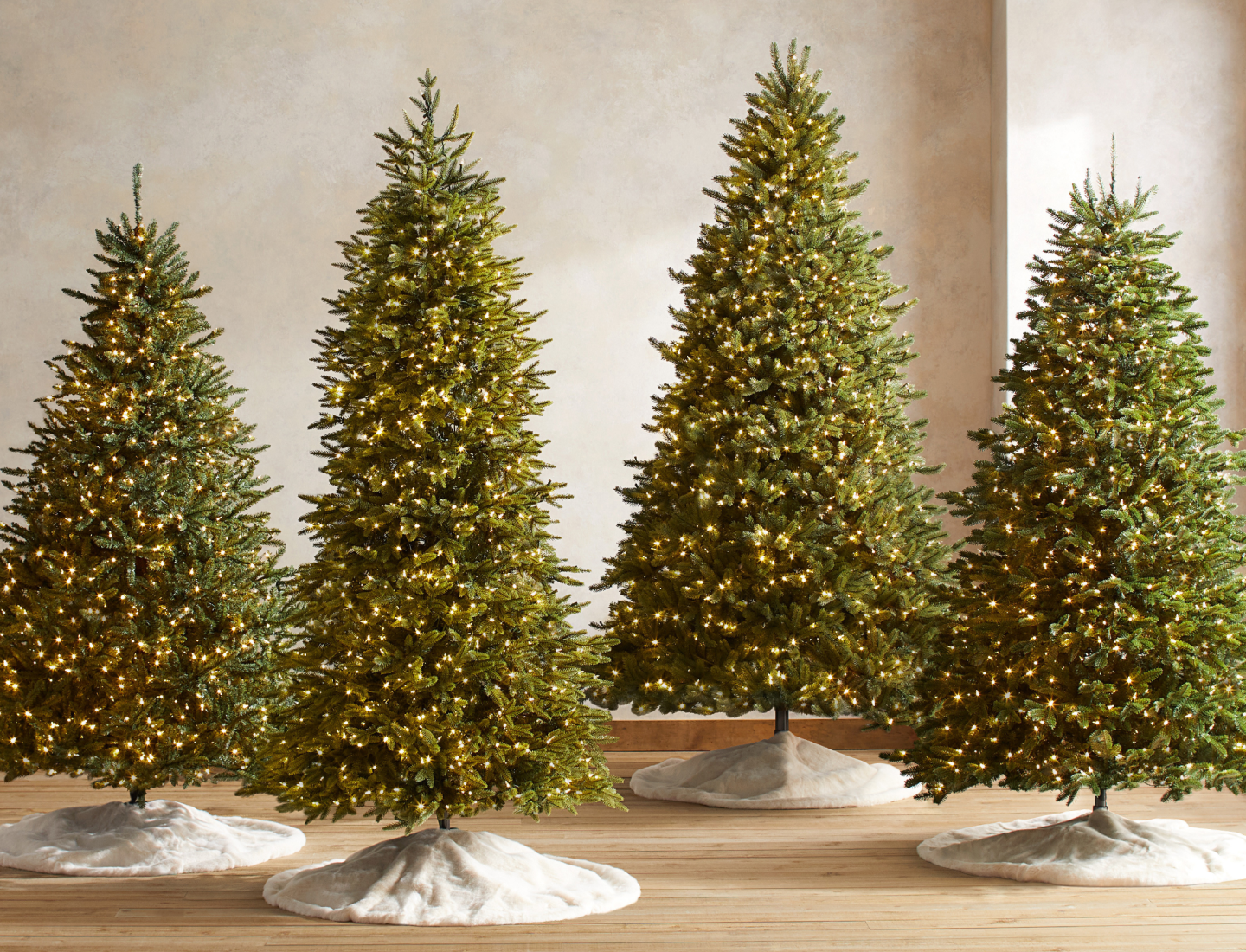 Group of four pre-lit artificial Christmas trees in different shapes and sizes, each displayed with a white tree skirt.