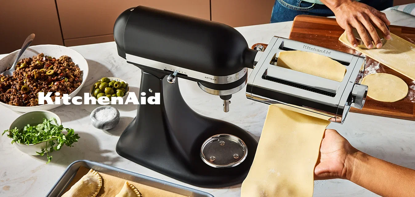Twee personen die pasta maken met een pastamachine, gevulde pasta op een bakplaat, kommen met vulling, olijven, kruiden en deeg op een keukentafel.