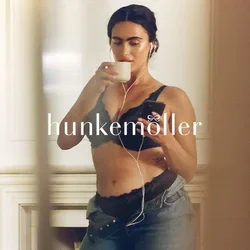 Zwarte kanten bralette met geschulpte randen, hooggetailleerde spijkerbroek en oortjes. Houdt met de ene hand een witte beker vast en met de andere een smartphone.