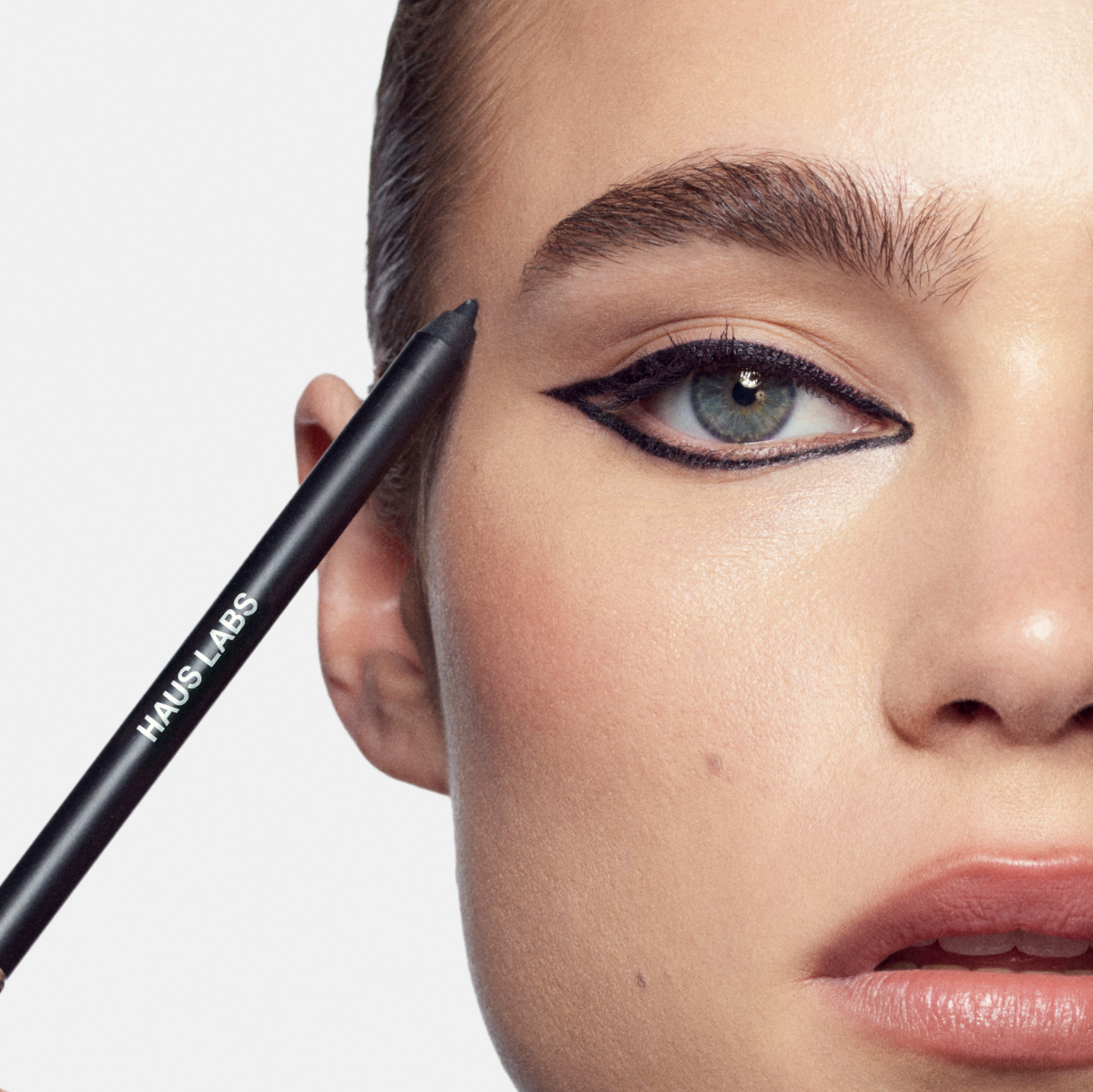 Close-up of a model with graphic black winged eyeliner, holding a Haus Labs eyeliner pencil beside the eye.