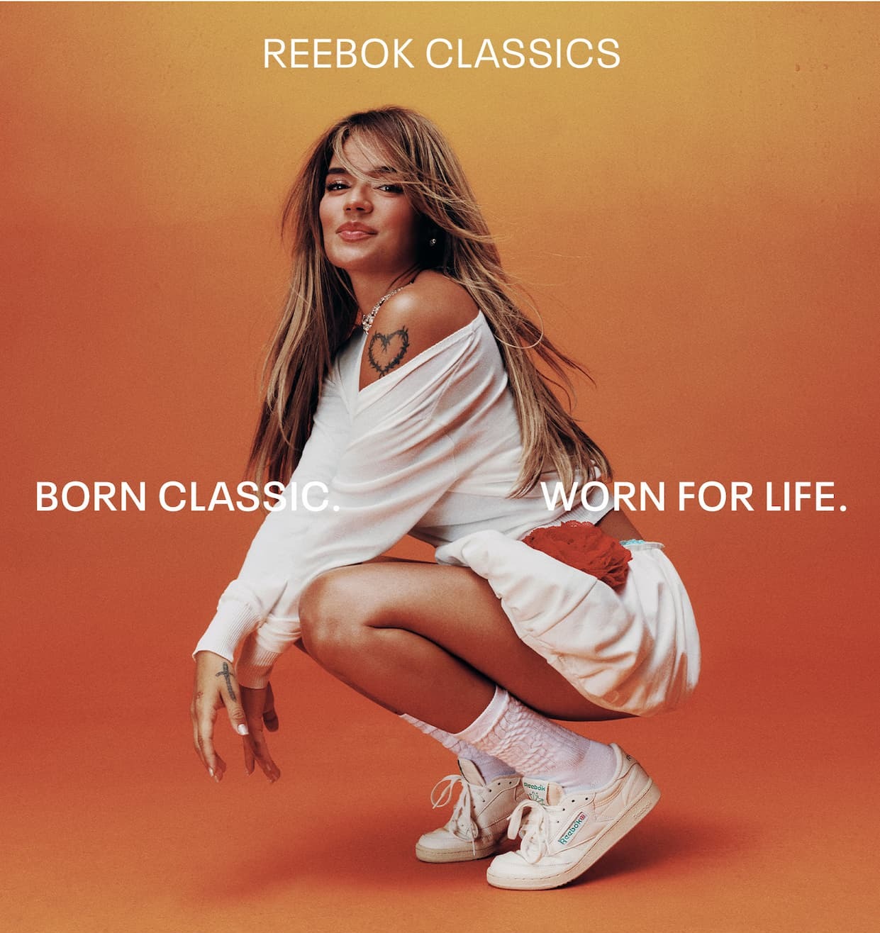 Person wearing white Reebok Classic sneakers crouches against an orange backdrop. Text reads: “REEBOK CLASSICS. BORN CLASSIC. WORN FOR LIFE.”
