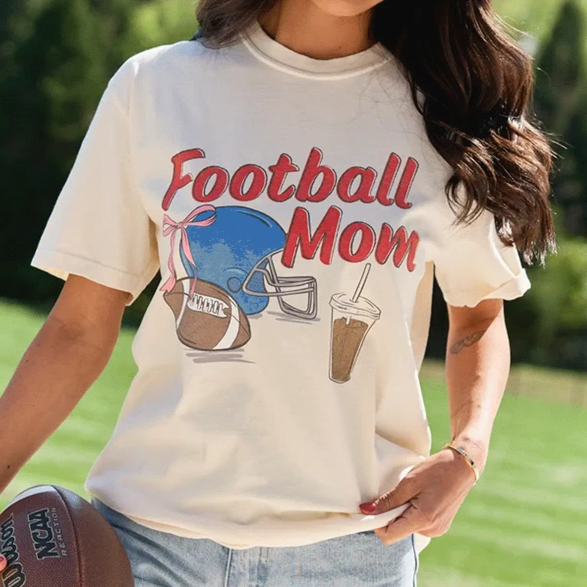 Woman wearing cream Football Mom graphic tee with blue helmet and football designs, standing outdoors holding a football.