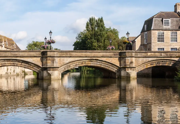 Stone bridge over a calm river, reflecting nearby buildings and trees under a clear sky.