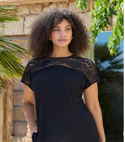 Ein Model trägt ein schwarzes Spitzenshirt mit Rundhalsausschnitt und transparentem Spitzeneinsatz in der Schulterpartie und eine weiße 7/8-Hose
