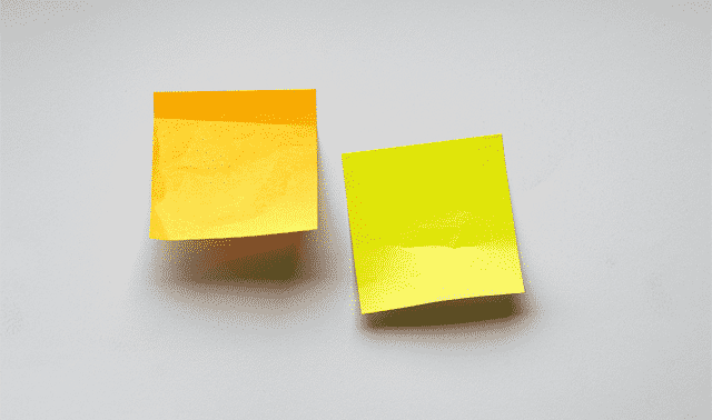 Two adhesive notes, one orange and one yellow, are stuck to a light gray wall. They are slightly curled at the edges.