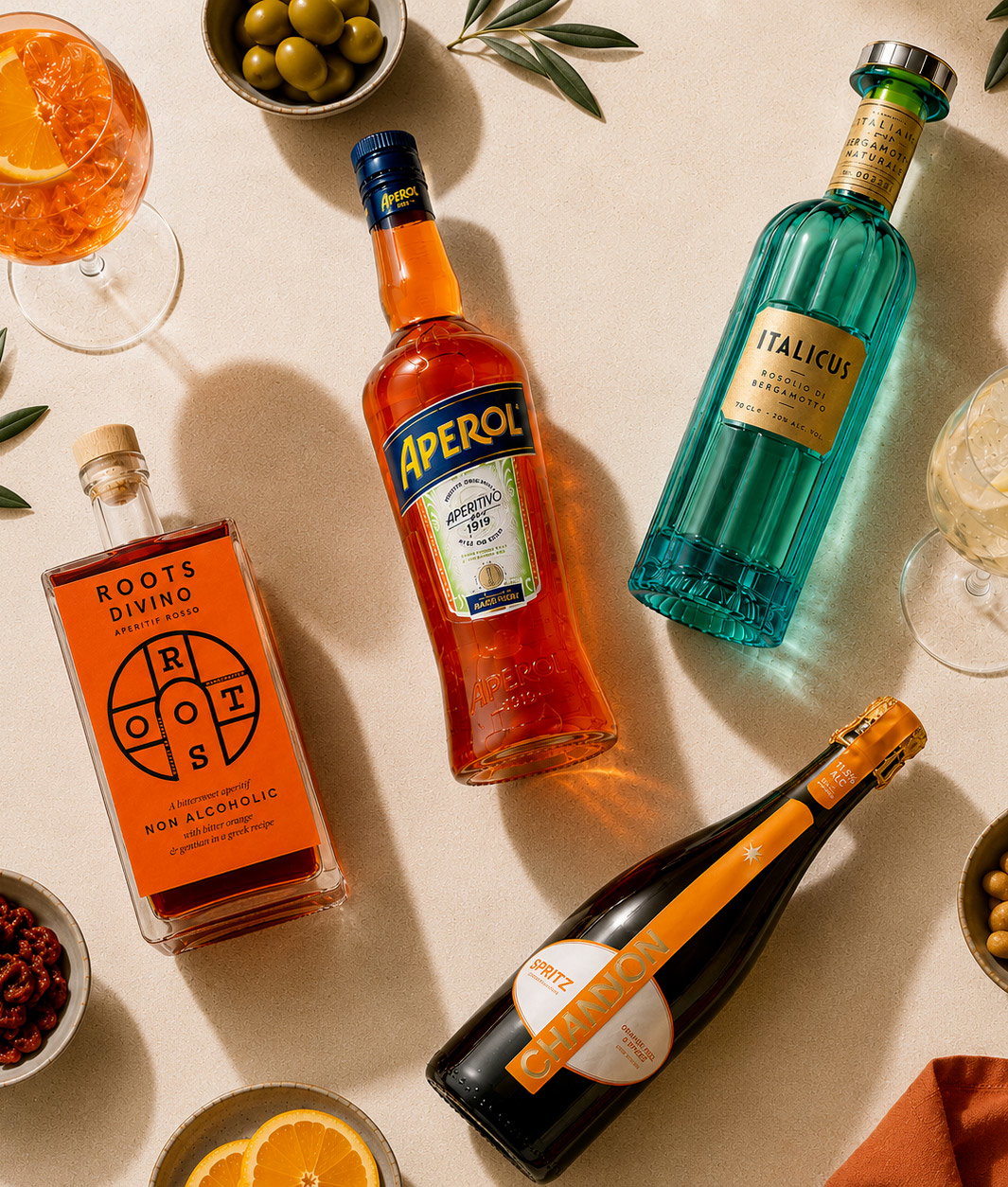 Overhead shot of Aperol, Italicus, Roots Divino non-alcoholic and Chandon bottles with spritz glasses, olives and orange garnish