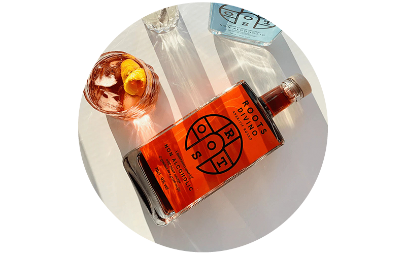 Bottle of Roots Divino non-alcoholic aperitif lying on its side with an iced cocktail glass garnished with citrus peel on a white surface