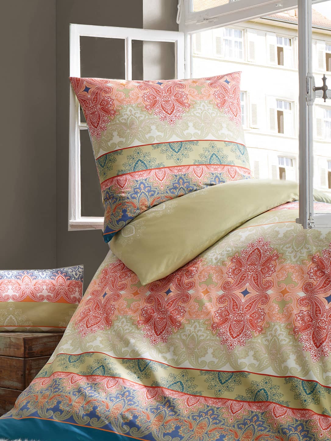 Ein Bett mit bunter Bettwäsche in Rot, Grün und Blau mit floralen und ornamentalen Mustern, dazu ein passendes Kissen, im Hintergrund ein geöffnetes Fenster mit Blick auf ein Gebäude.