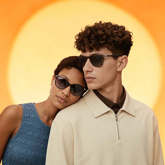 A person wearing a blue sleeveless top and sunglasses leans against another person in a beige zip-up shirt and sunglasses. They stand in front of a bright orange and yellow background resembling a sunset, creating a warm and stylish atmosphere.