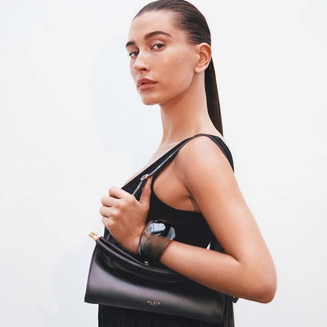 A person with sleek, dark hair pulled back holds a black Alaïa handbag against a plain background. They are wearing a sleeveless black dress and have a neutral expression.