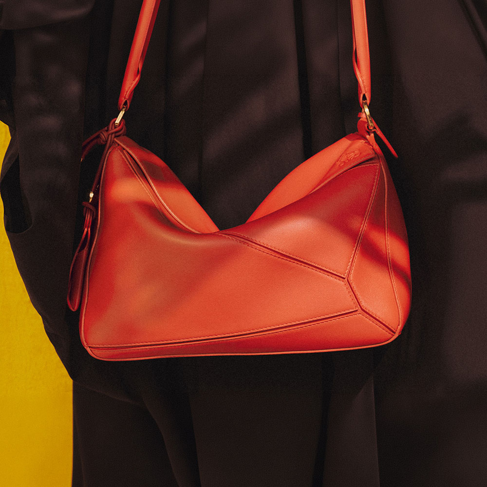 A stylish, geometric-shaped red handbag with a long strap, worn against a black outfit. The background features a bright yellow surface, creating a striking contrast.