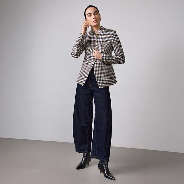 A person stands against a plain backdrop, wearing a checked blazer, dark wide-leg trousers, and black ankle boots. Their hair is pulled back, and they are adjusting one earring. The outfit is smart-casual with a modern touch.