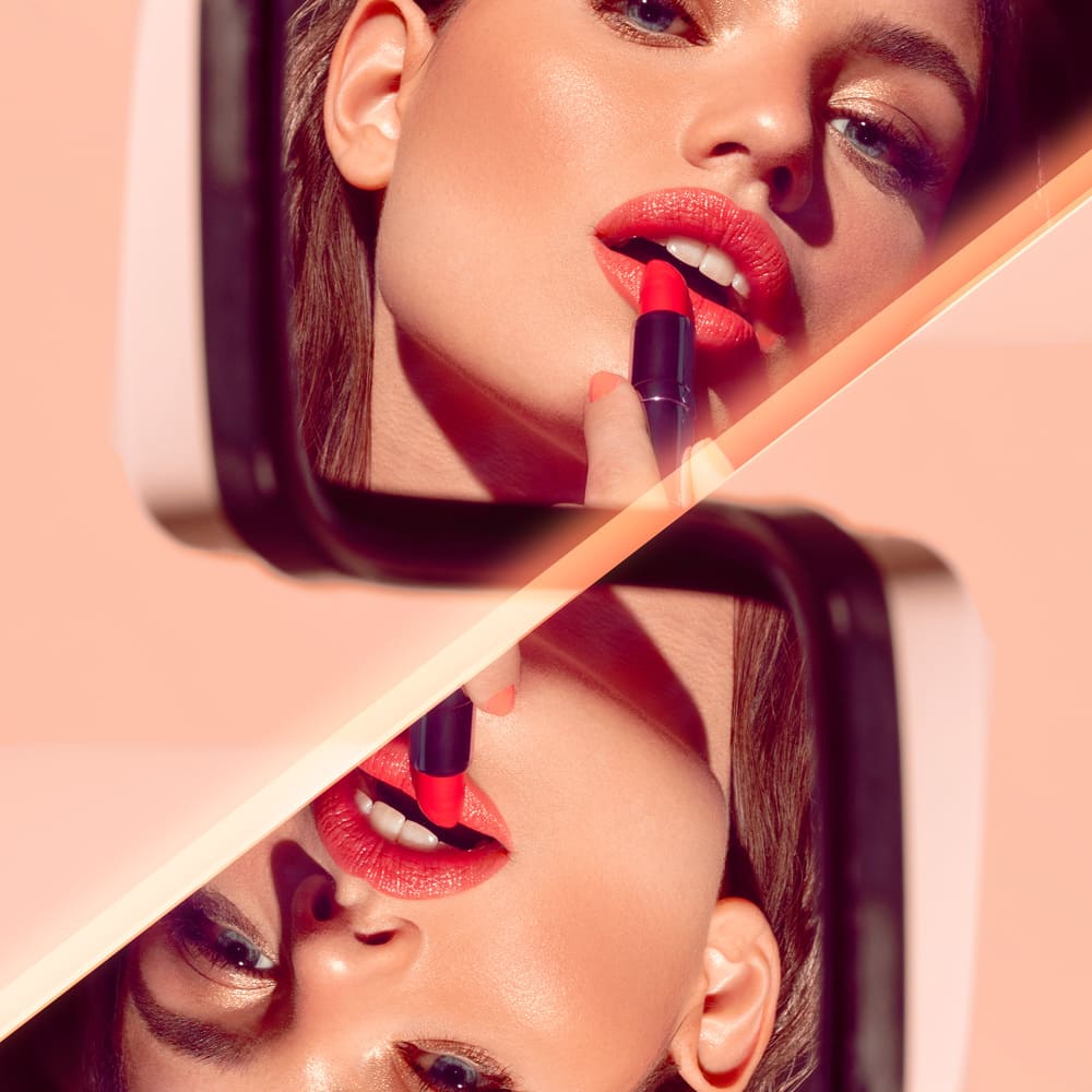 Close-up of woman applying red lipstick with a split mirrored reflection against a peach background, glossy makeup