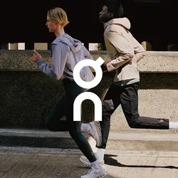Twee mensen joggen buiten met sportjacks, leggings en hardloopschoenen op een zonovergoten stadsstraat.
