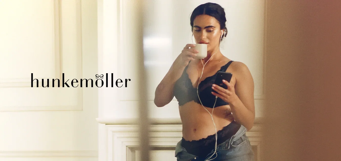 Zwarte kanten bralette met geschulpte randen, hoogwater denim jeans en oordopjes. Houdt met de ene hand een witte beker vast en met de andere een smartphone.