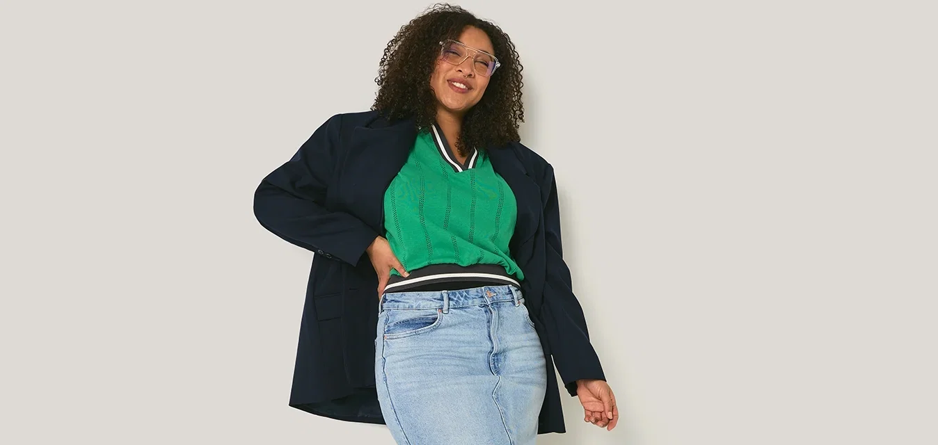 Jonge vrouw met krullend haar lacht, draagt een groene top, spijkerrok, zwarte jas en zwarte laarzen, staand tegen een effen lichte muur.