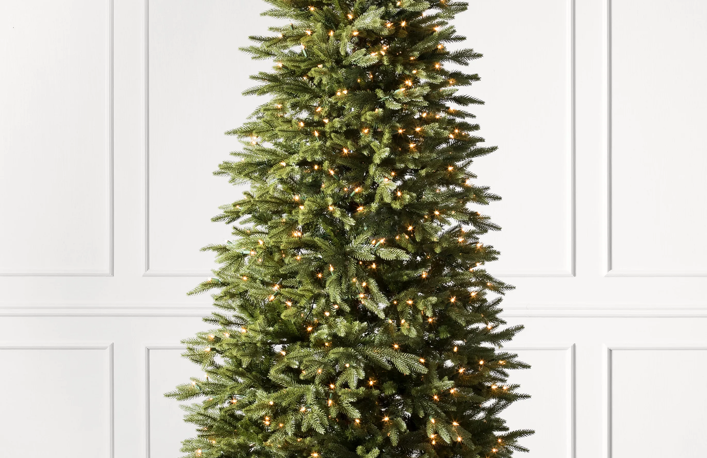 Pre-lit artificial Christmas tree with layered green branches and warm white lights against a white panelled wall.