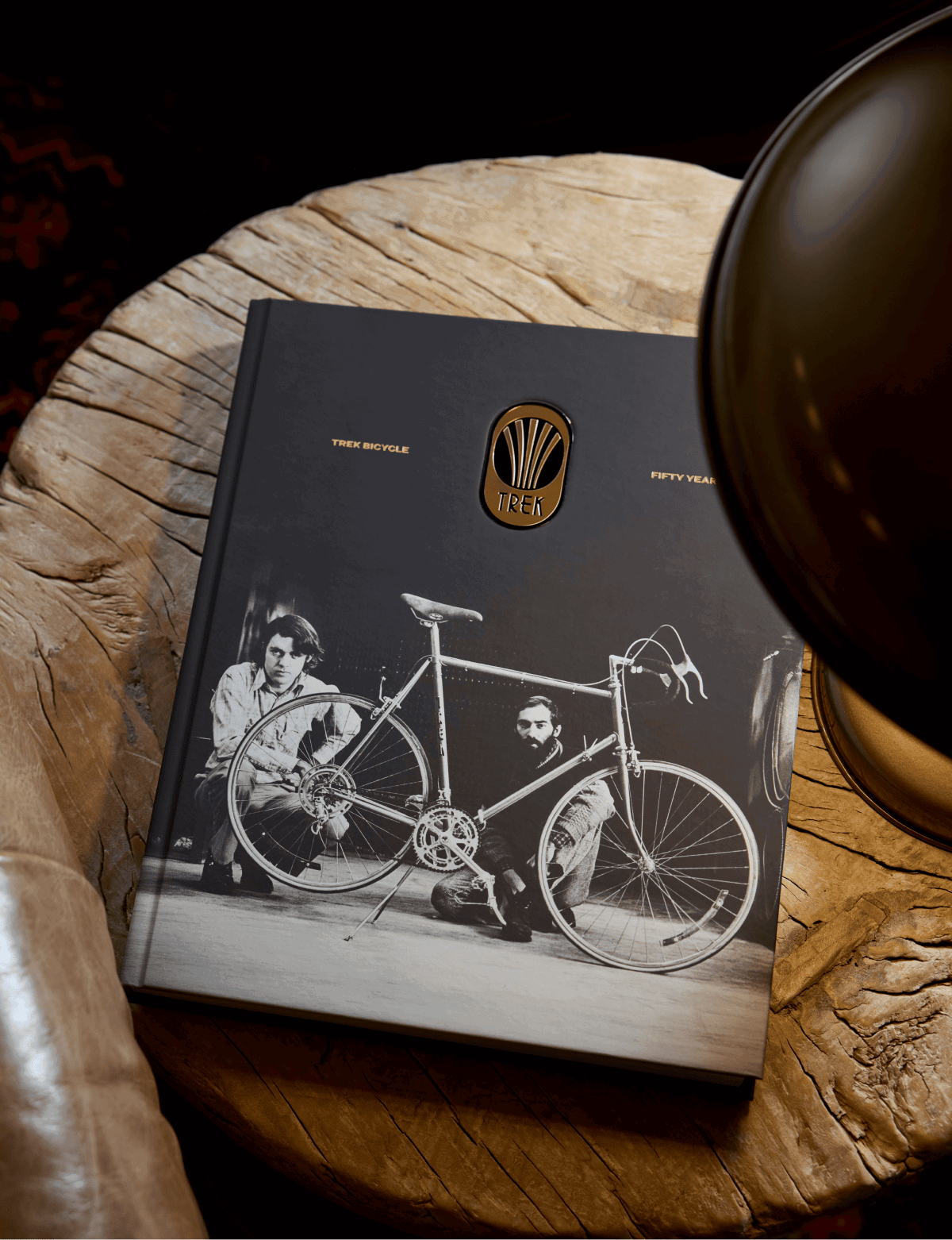 The Trek 50th Anniversary Book, a black hardcover with a black-and-white photo of two people and an early Trek bicycle, resting on a wood table