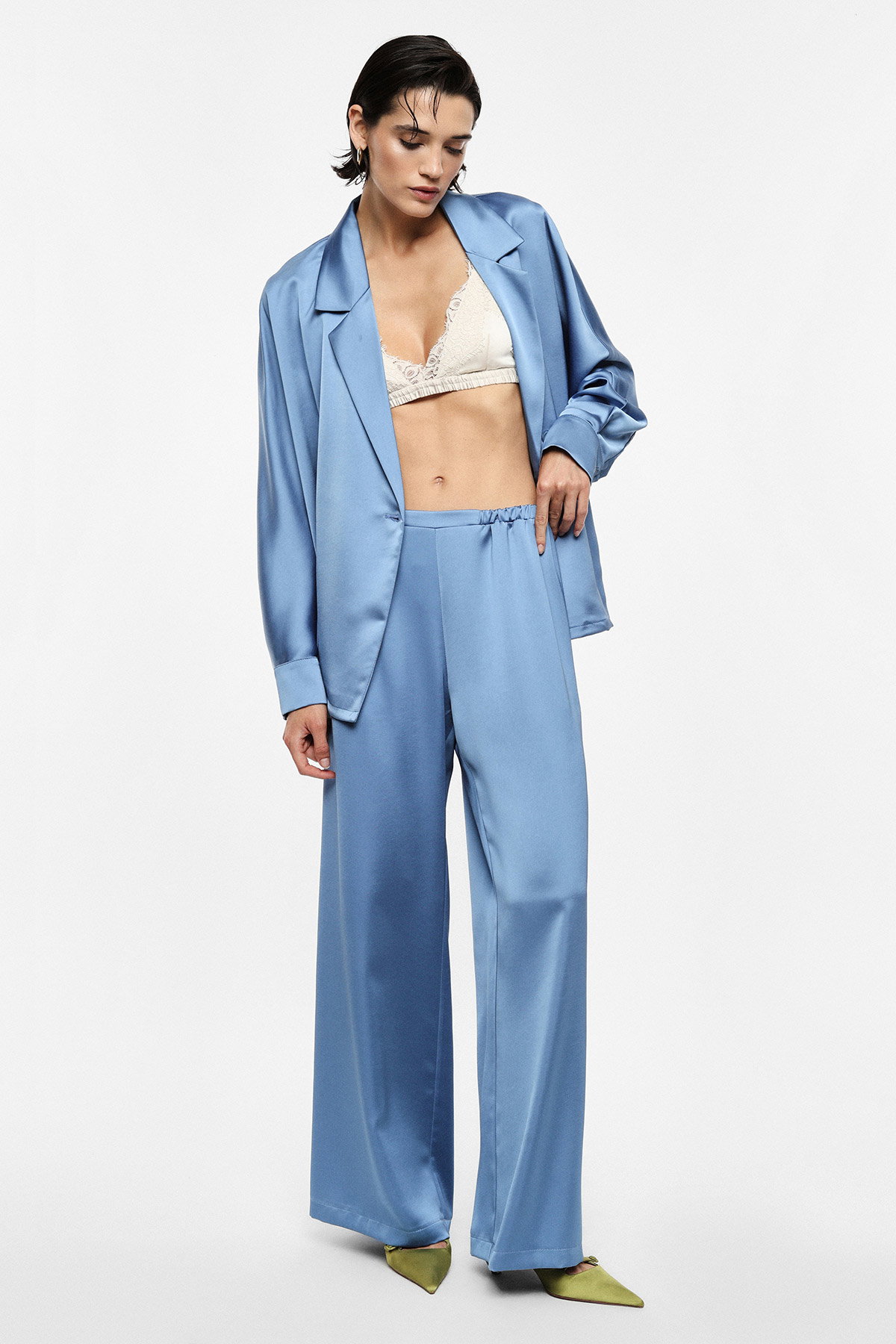 Model wearing light-blue satin oversized blazer and wide-leg trousers with cream lace bralette and green pointed heels on white background