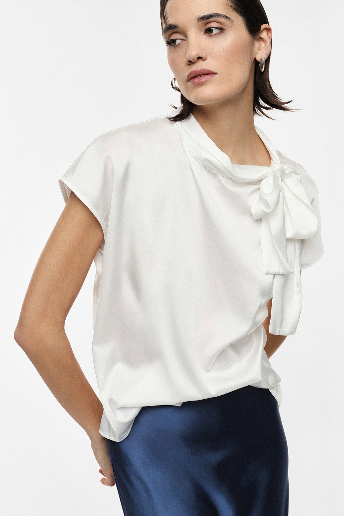 Woman wearing a white silk bow-tie blouse and navy satin skirt, short dark hair and hoop earrings, three-quarter pose