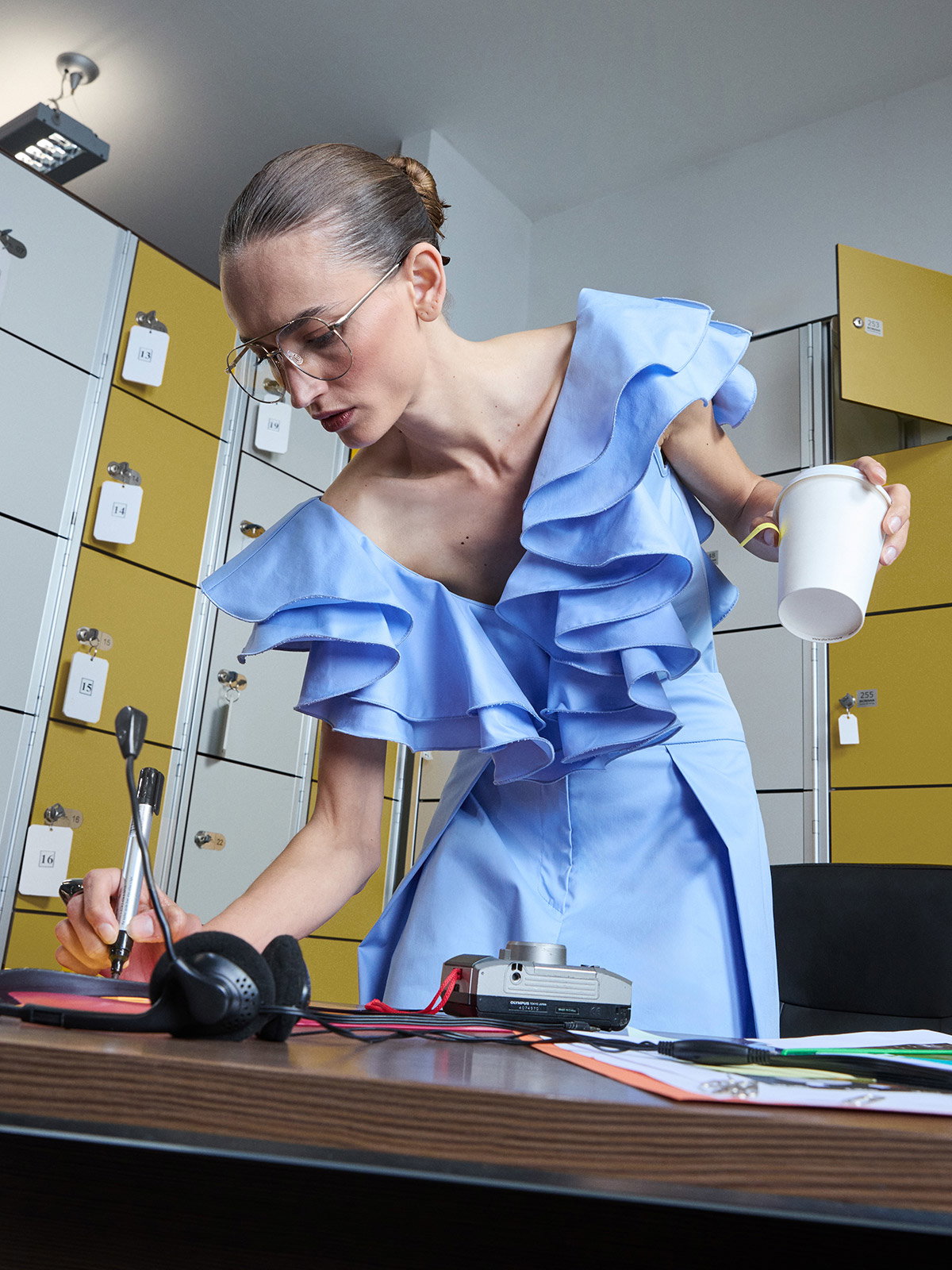 Woman in blue ruffled dress leaning over a desk in a locker room, holding a takeaway cup and writing on paper