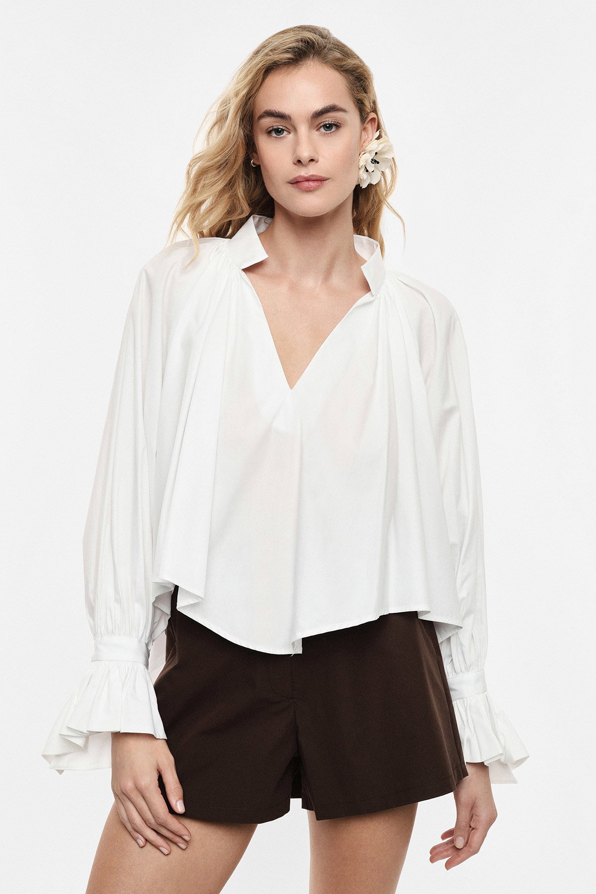 Model wearing loose white V-neck blouse with ruffled cuffs, brown tailored shorts and a white flower hair accessory on plain background