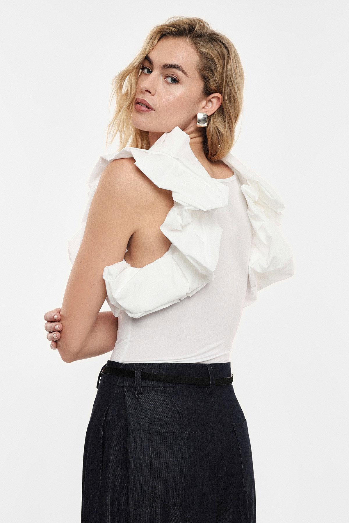 Model wearing white ruffle sleeveless top and black trousers, looking over her shoulder