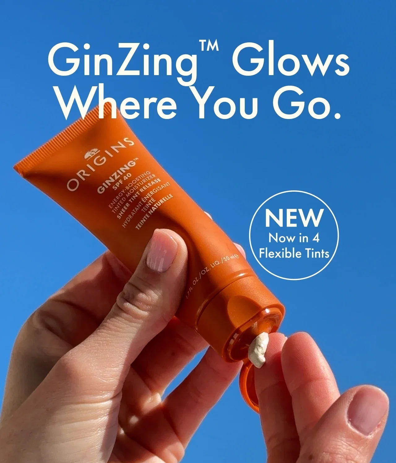 Hand squeezes cream out of a tube of GinZing™ SPF 40 Tinted Moisturizer, with text above reading, 'GinZing™ Glows Where You Go', against a sky blue background.