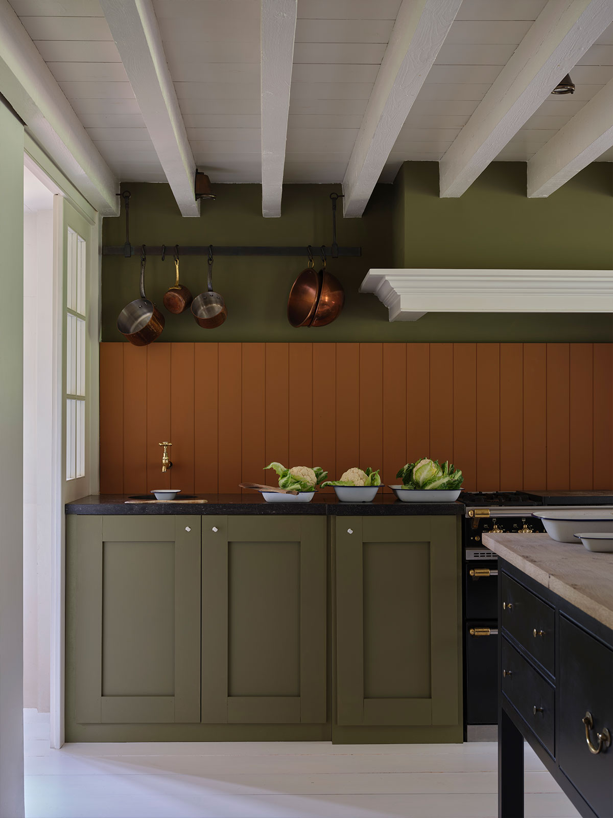 Olive and terracotta kitchen with copper pans, white beams, and dark worktops.